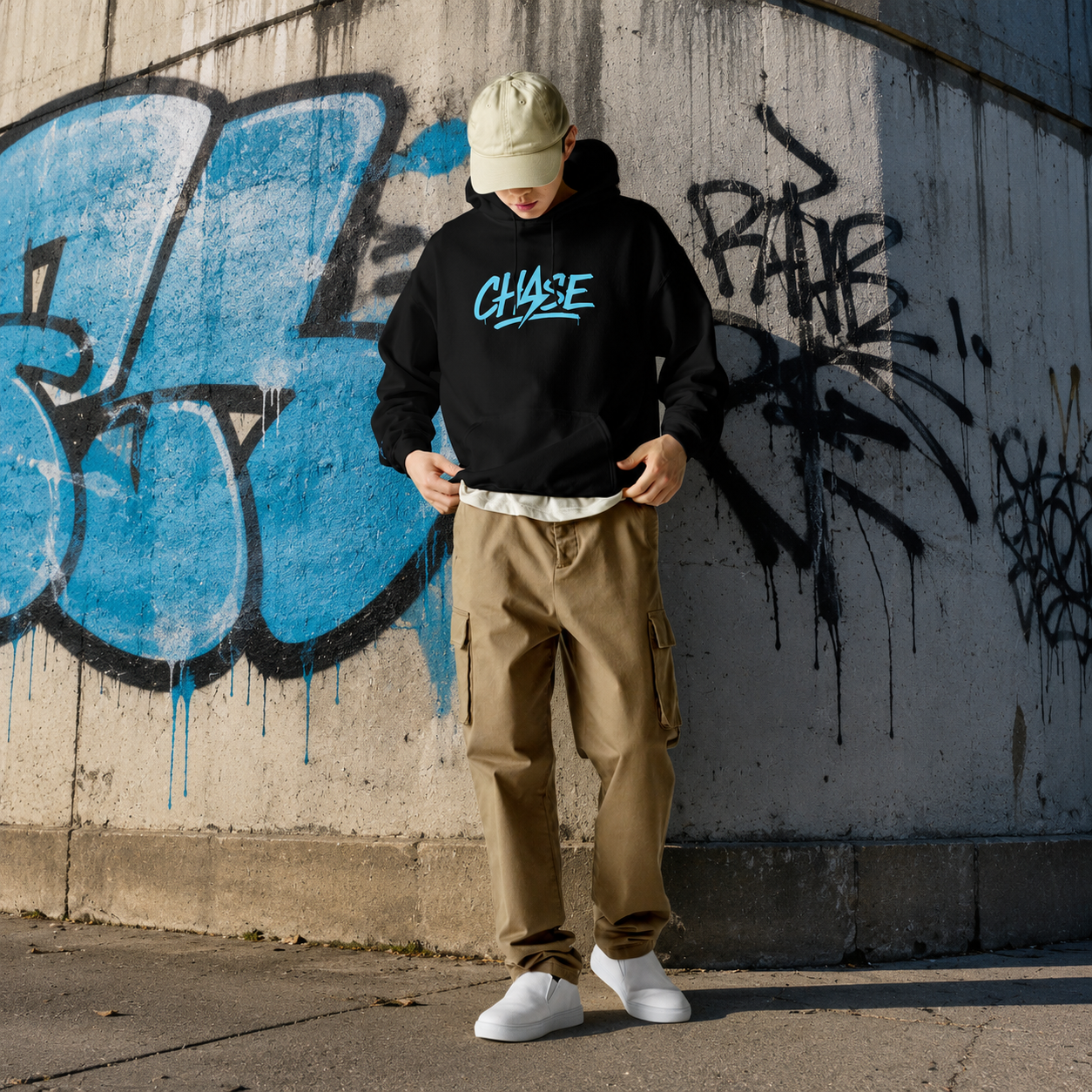 Model wearing the black CHASE Core Hoodie in front of a blue graffiti wall